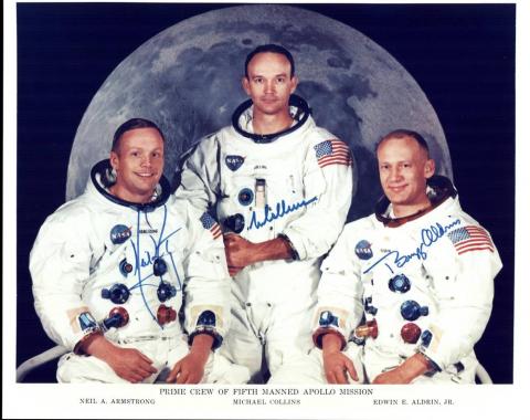 The Apollo 11 crew in their spacesuits but not wearing space helmets. They are shown in front of a large image of the Moon. Neil Armstrong is seated on the left. Michael Collins is standing in the centre. Buzz Aldrin is seated on the right.